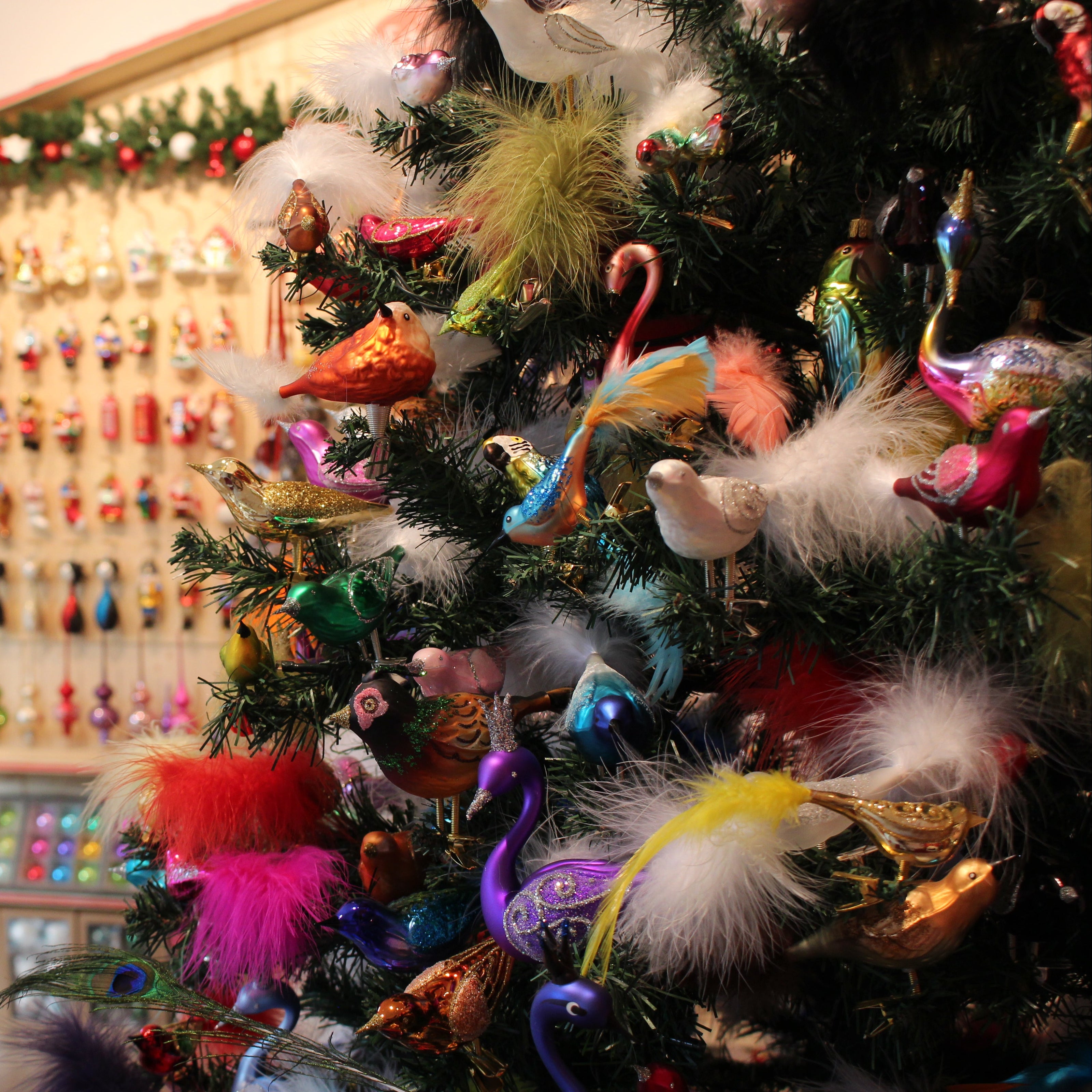 A close-up of a decorated Christmas tree with colorful ornaments, white feathers, and metallic accents, set in a shop interior with shelves of holiday decorations in the background.